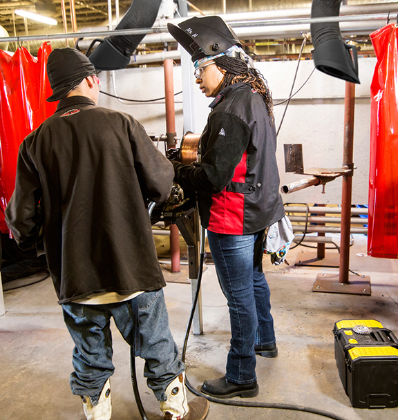 Two welding students in a lab standing by a MIG welding machine with wire feeder